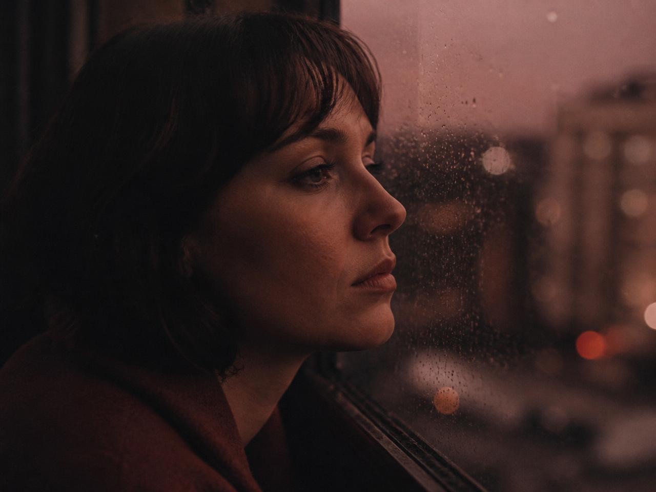 A woman in profile looking out a rain-streaked window at dusk